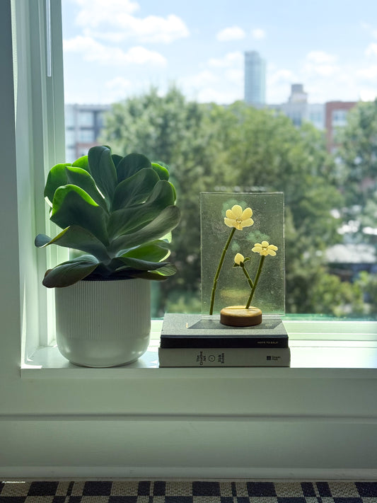 A glass memorial suncatcher with the ashes and cremains embedded into the pistol of the flower sitting on a wood stand in a widow with the light pouring through it