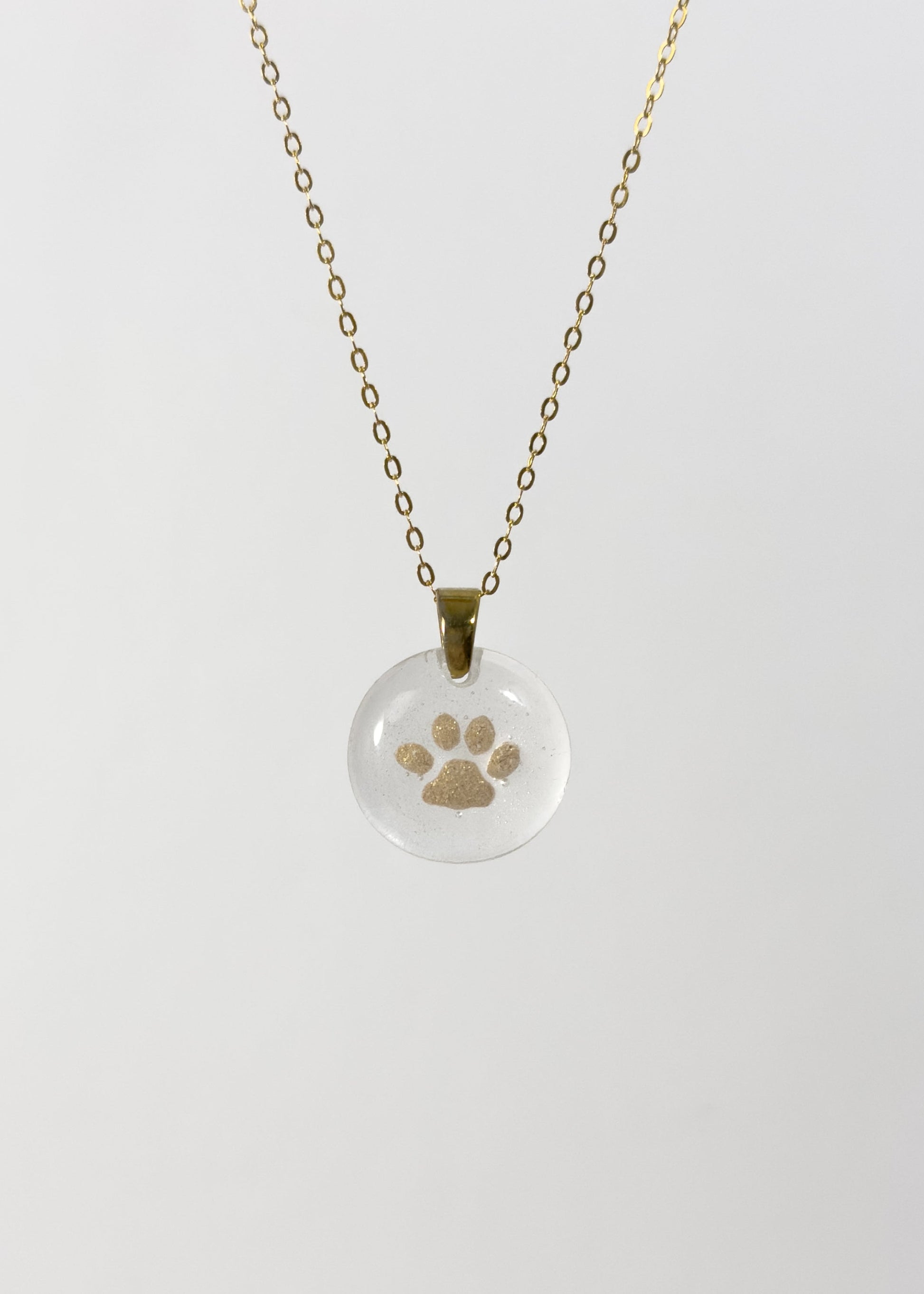 Clear glass pendant necklace with a gold cat paw print design made out of cremains on a white background