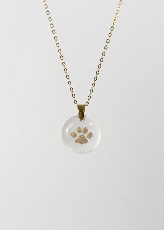 Clear glass pendant necklace with a gold cat paw print design made out of cremains on a white background