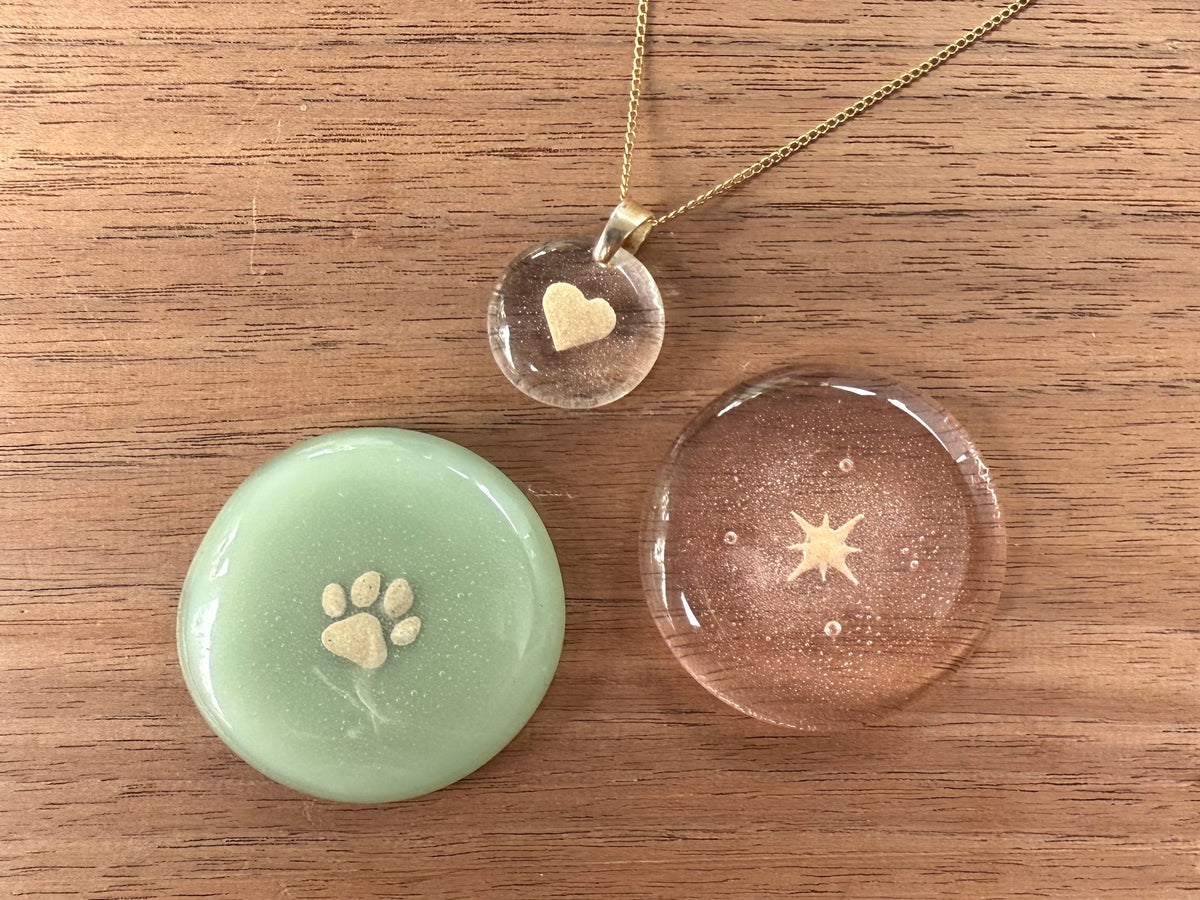 One touchstone with a paw print, a touchstone with ashes shaped into a star and  a pendant with ashes shaped into a heart, sitting on a wooden surface.