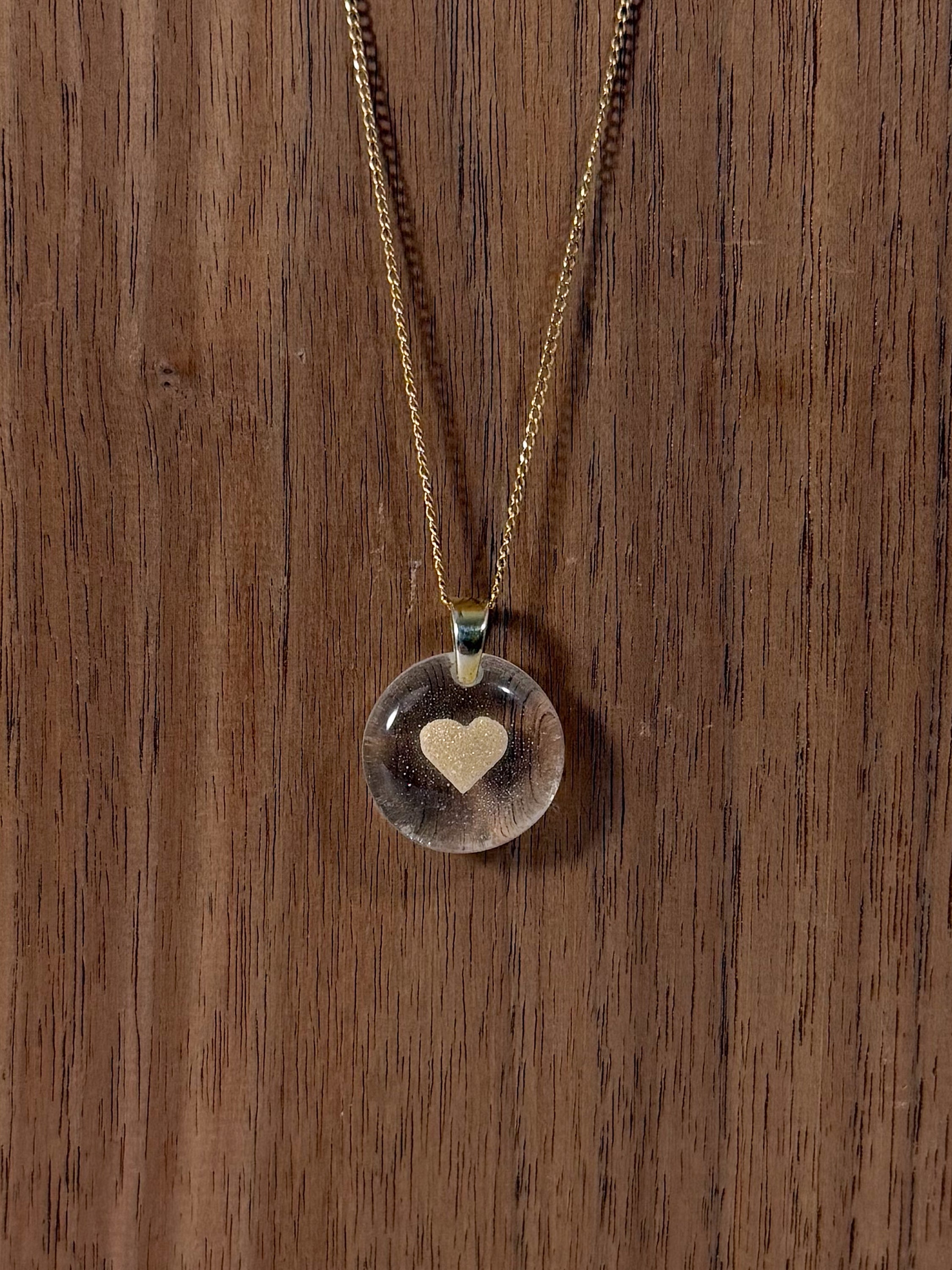 A clear glass pendant with the cremains shaped into a heart floating in the center, the necklace is gold.