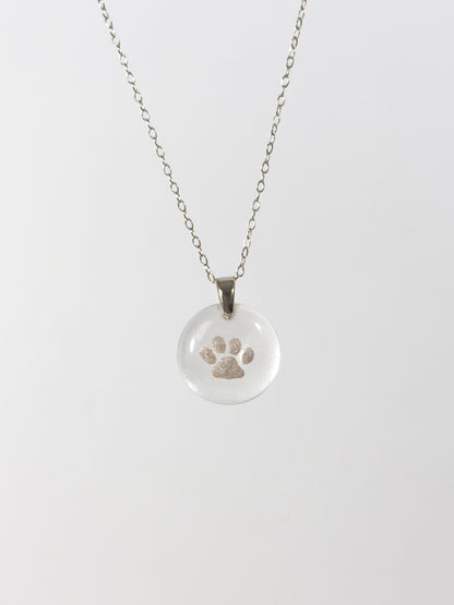 Clear glass pendant necklace with a silver cat paw print design made out of cremains on a white background